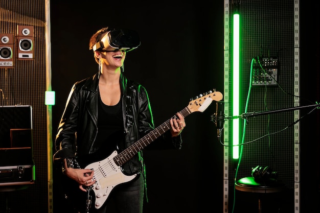 Guitarrista com fone de ouvido de realidade virtual desfrutando de simulação de concerto de rock enquanto toca música de heavy metal na guitarra elétrica no estúdio de música. Músico de mulher tocando álbum grunge usando instrumento elétrico