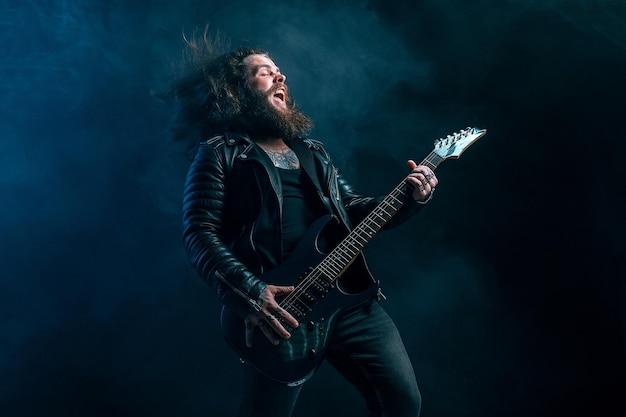 Guitarrista masculino de rock expressivo com cabelo comprido e barba toca no tiro de estúdio de fundo de fumaça