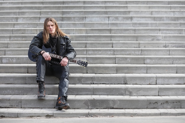 Guitarrista de rock en los escalones. Un músico con un bajo en un traje de cuero. Metalista con una guitarra en el fondo del paso industrial.