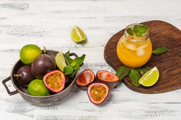 Gulupa o jugo de maracuyá rodeado de cáscaras y frutas enteras y cortadas, limones y menta verde sobre fondo blanco de madera envejecida