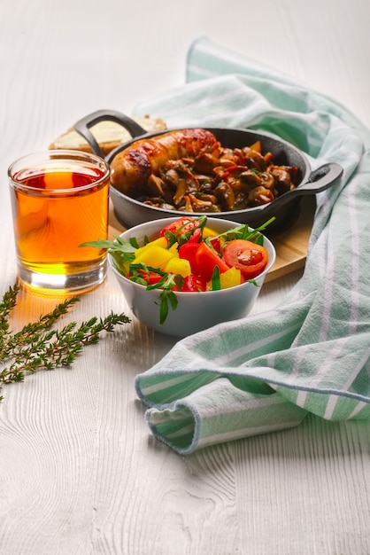 Gusseisenpfanne mit gebratener wurst und pilzen, salat mit frischgemüse und glas saft auf weißem holztisch.