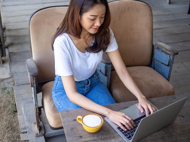 Gut aussehende asiatische Frauen verwenden Computer, um eine Verbindung zum Internet herzustellen, um mit ausländischen Freunden zu kommunizieren Eine glückliche und entspannte Frau sitzt auf dem Sofa Arbeitsfrau und freiberufliches Konzept