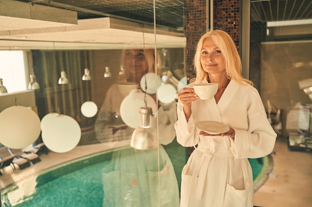 Foto gut aussehende frau mit einer tasse kaffee in der hand, während sie im resorthotel vor der kamera posiert?