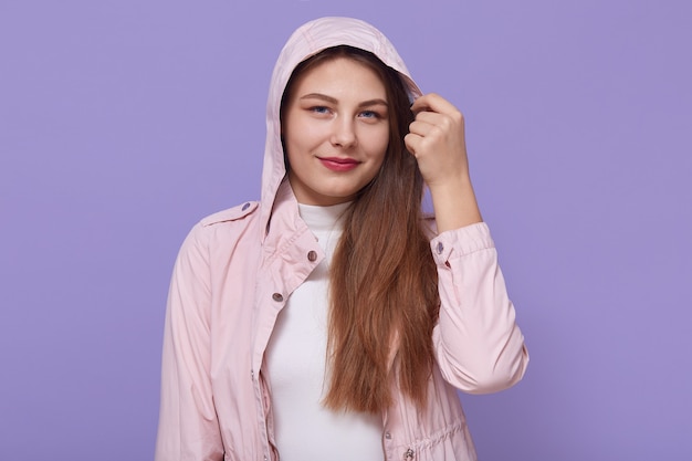 Gut aussehende Frau mit schönen Augen, die blassrosa Jacke mit Kapuze tragen, ihren Kopfschmuck berührend, mit ruhigem Lächeln gegen lila Hintergrund stehend.