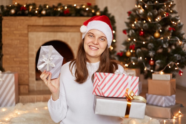 Gut aussehende, glückliche, optimistische Frau mit weißem Pullover und rotem Weihnachtsmütze, die viele Geschenkboxen in den Händen hält, Silvester feiert, in der Nähe von Kamin und Weihnachtsbaum posiert.