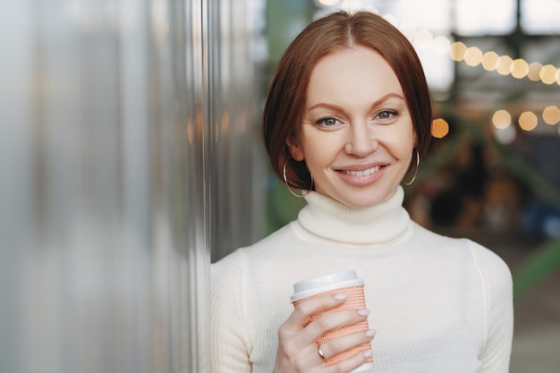 Gut aussehende junge Frau in einem lässigen weißen Rollkragenpullover hält eine Papptasse mit aromatischem Cappuccino oder Kaffee, die glücklich auf die Kamera posiert und draußen ein Freizeit-Lifestyle-Konzept hat