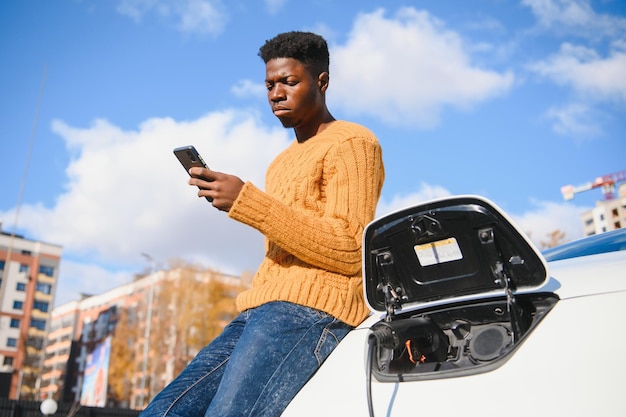 Gut aussehender afrikanischer Mann, der in der Nähe von Luxus-Elektroautos steht und auf dem Handy spricht.