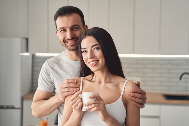 Gut aussehender bärtiger Mann, der sanft die Schultern seiner jungen Frau umarmt. Hübsche Frau, die eine Tasse Tee in den Händen hält