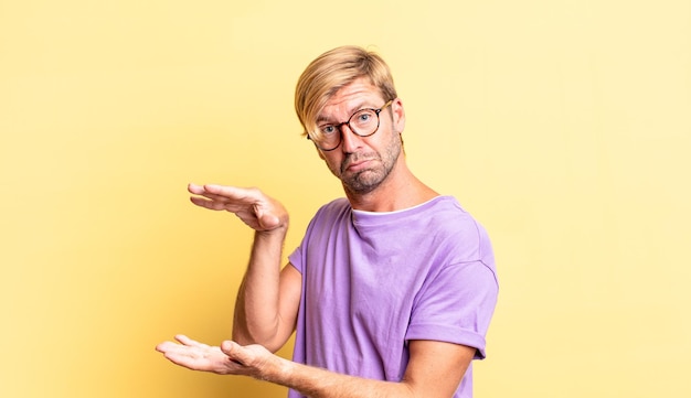 Foto gut aussehender blonder erwachsener mann, der ein objekt mit beiden händen auf der seite des kopierraums hält, ein objekt zeigt, anbietet oder annonciert