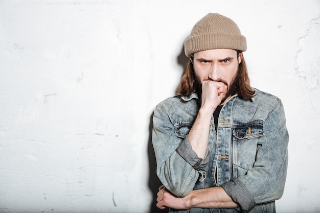 gut aussehender ernster bärtiger Hipster-Mann mit Hut in Jeansjacke, der über der Wand posiert