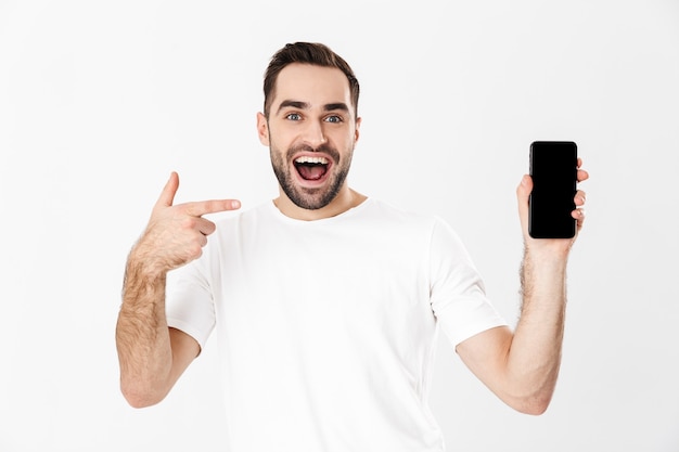 Gut aussehender fröhlicher Mann mit leerem T-Shirt, das isoliert über weißer Wand steht und leeres Handy zeigt