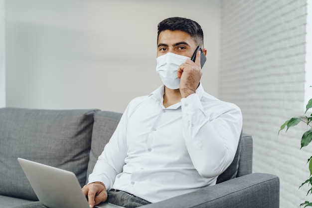 Gut aussehender Geschäftsmann des Mannes in der medizinischen Maske, die seinen Laptop benutzt, während er auf seinem Handy drinnen spricht