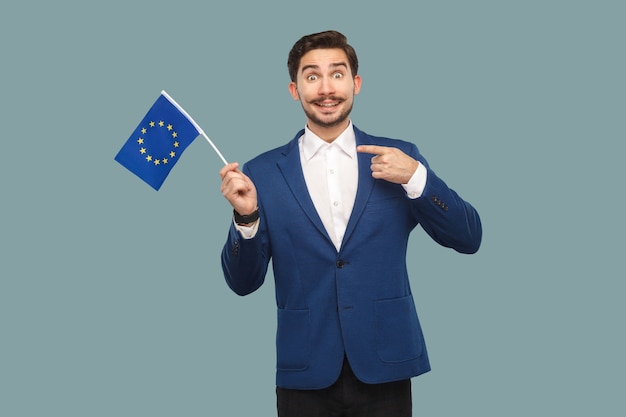Gut aussehender Geschäftsmann in blauer Jacke und weißem Hemd, der mit dem Finger auf die Flagge der Europäischen Union zeigt und mit einem zahnigen Lächeln in die Kamera schaut. Innen, Studioaufnahme auf hellblauem Hintergrund isoliert.