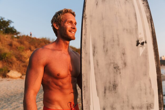 gut aussehender hemdloser Surfer, der bei Sonnenaufgang sein Surfbrett am Meer hält