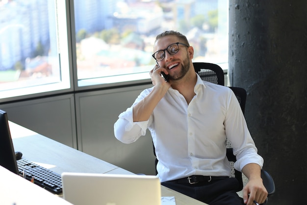 Gut aussehender junger Geschäftsmann im Hemd, der am Smartphone spricht und lächelt, während er im Büro sitzt.