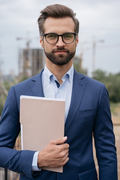 Foto gut aussehender manager mit anzug und brille, der geschäftsdokumente mit blick in die kamera hält