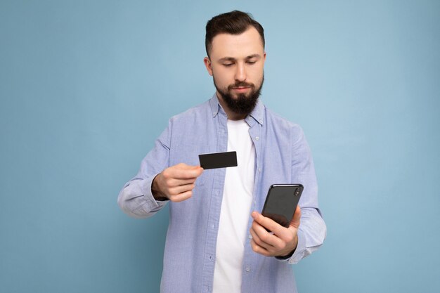 Gut aussehender Mann, der Alltagskleidung trägt, isoliert auf der Hintergrundwand, die Telefon und Kreditkarte hält und die Zahlung mit Blick auf den Smartphone-Bildschirm macht