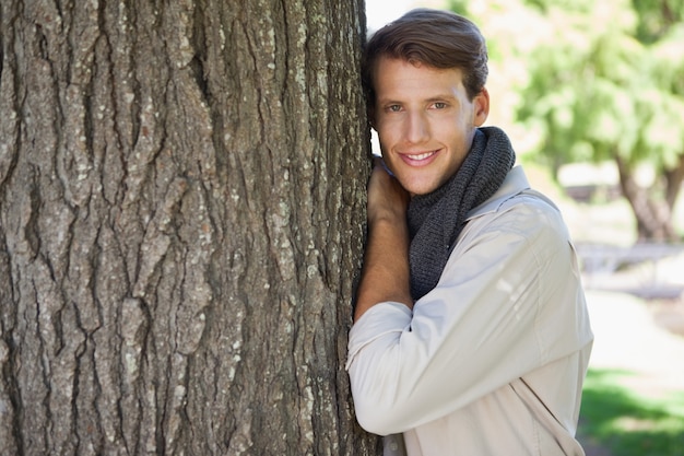 Gut aussehender Mann, der am Baum lächelt an der Kamera sich lehnt