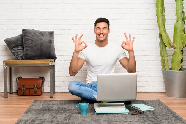 Gut aussehender Mann, der auf dem Boden mit seinem Laptop zeigt ein okayzeichen mit den Fingern sitzt