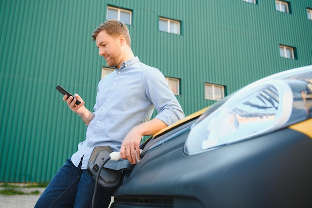 Gut aussehender Mann, der das Telefon benutzt, während das Auto aufgeladen wird