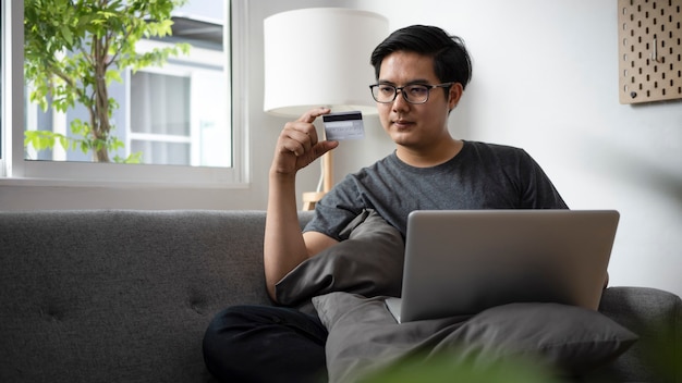 Gut aussehender Mann, der Kreditkarte hält und Laptop-Computer im Wohnzimmer verwendet.