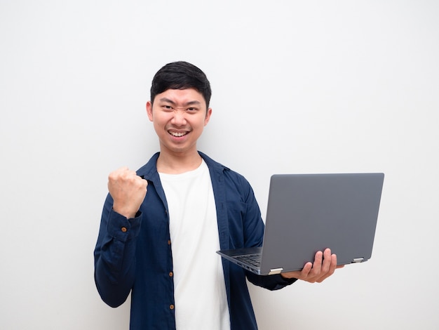 Gut aussehender Mann, der Laptop hält, zeigt die Faust nach oben, selbstbewusstes Gesicht, Blick in die Kamera auf weißem Hintergrund