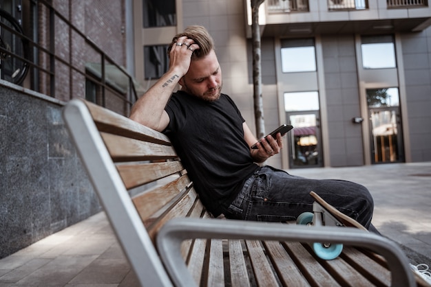 Gut aussehender Mann, der mit dem Smartphone im Internet surft, Beiträge mag und sie über soziale Netzwerke teilt, Nachrichten auf der Bank mit Longboard mit nachdenklichem Blick in schwarzer Jeans und T-Shirt liest