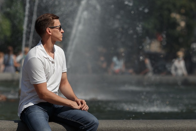 Gut aussehender Mann, der mit Sonnenbrille auf dem Brunnenhintergrund sitzt, weg von der Kamera. Exemplar