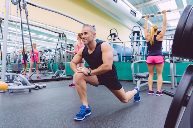 Gut aussehender Mann, der sich in einem Fitnesscenter ausdehnt, und zwei schöne Frauen, die im Hintergrund Übungen mit Hanteln machen