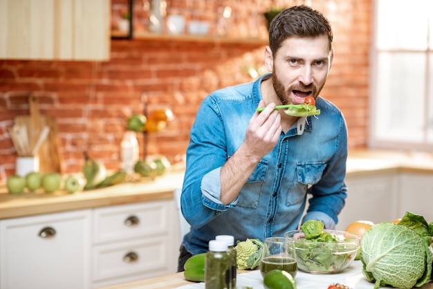 Gut aussehender Mann, der zu Hause gesunden Salat isst, der am Tisch voller grüner Zutaten in der Küche sitzt