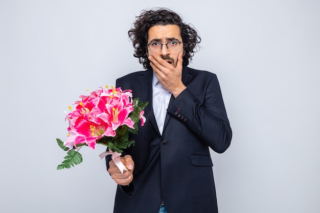 Gut aussehender Mann im Anzug mit Blumenstrauß, der schockiert aussieht und den Mund mit der Hand bedeckt