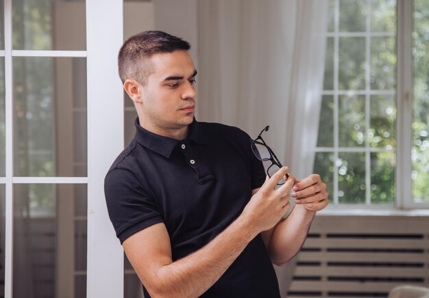 gut aussehender mann in einem schwarzen t-shirt betrachtet eine stilvolle brille mit einem schwarzen rand in seinen händen im raum auf dem hintergrund des fensters. Nahansicht