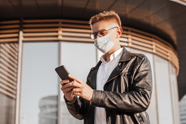 Gut aussehender Mann in einer Schutzmaske im Gesicht mit Brille mit einem Smartphone auf der Straße einer Großstadt. Geschäftsmann, der auf städtischem Hintergrund telefoniert