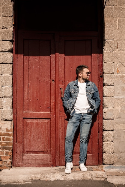 Gut aussehender Mann mit Bart in Jeansjacke und Jeanshose steht in der Nähe von roten Vintage-Türen. Erwachsener Typ im Denim-Style