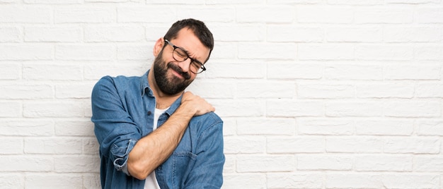 Gut aussehender Mann mit Bart über der weißen Backsteinmauer, die unter den Schmerz in der Schulter für das Haben einer Bemühung leidet