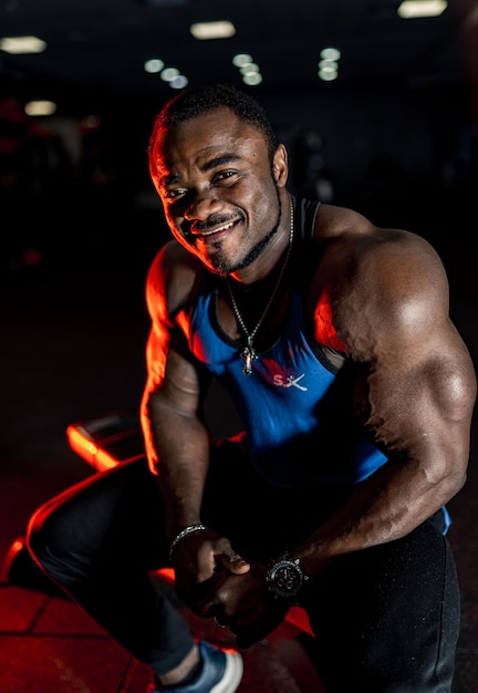 Gut aussehender Mann mit großen Muskeln, der in einem Fitnessstudio vor der Kamera posiert. Dunkler heller Hintergrund. Porträt eines lächelnden Bodybuilders. Nahansicht.