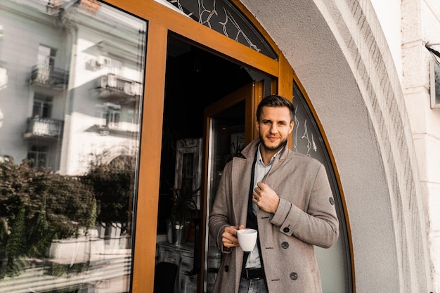 Gut aussehender Mann mit Tasse Kaffee posiert im Mantel im Café. Männlicher Lebensstil.