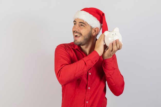 Gut aussehender Mann mit Weihnachtsmütze, der ein Sparschwein auf weißem Hintergrund hält Sparkonzept zum Jahresende