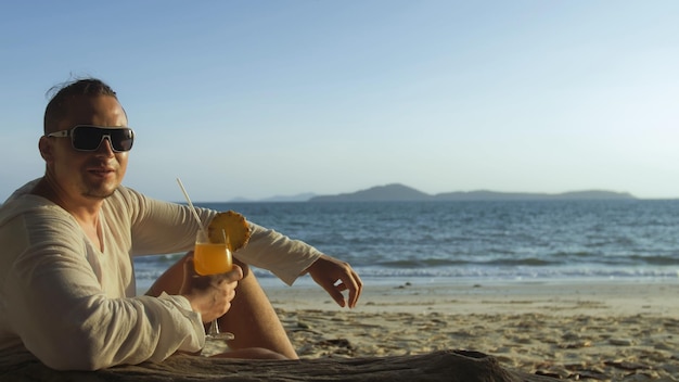 Gut aussehender Mann sitzt in weißem Kleid Sonnenbrille trinken Ananas-Cocktail Pina Colada Schöner Strand Meereswellen bei Sonnenuntergang Konzept Ruhe tropisches Resort Reisen Tourismus glückliche Sommerferien