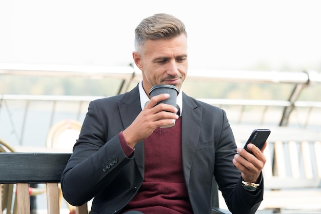 Gut aussehender Mann trinkt Kaffee, der morgens im Freien Wirtschaftsnachrichten auf dem mobilen Gerät liest und trinkt.