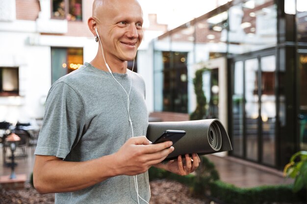 Gut aussehender positiver junger Yoga-Mann, der im Freien Fitness-Teppich hält und Musik mit Kopfhörern auf dem Handy hört.