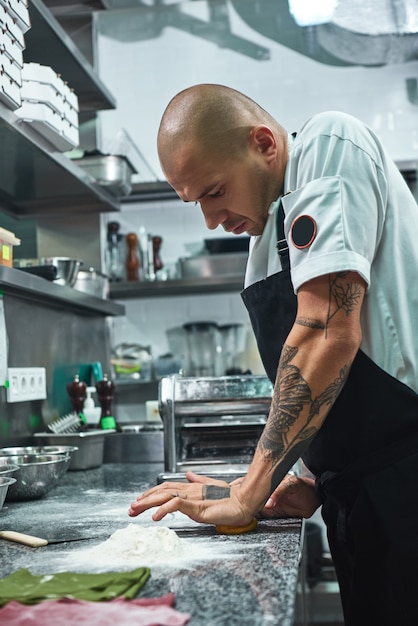 Gut aussehender Profikoch mit Tätowierungen an den Händen, der den Teig in der Restaurantküche knetet