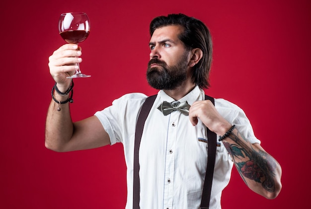 Foto gut aussehendes alkoholisches getränk, stilvoller sommelier, der altmodisches bärtiges hipster-weinmacher, trendiger mann in hosenträgern mit wein, selbstbewusster, eleganter mann, der wein aus glas trinkt