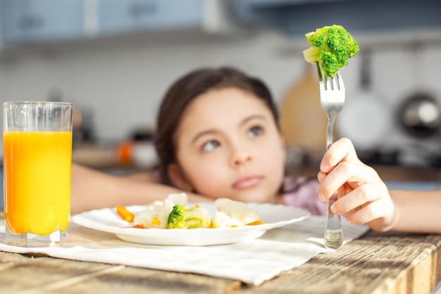 Gut für die Gesundheit. Ziemlich konzentriertes dunkelhaariges kleines Mädchen, das gesundes Frühstück hat und etwas Saft trinkt und das grüne Gemüse auf ihrer Gabel betrachtet