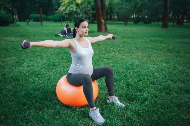 Gut gebaute junge schwangere Frau sitzen auf orange Fitnessball und trainieren im Park. Sie hält Hanteln.