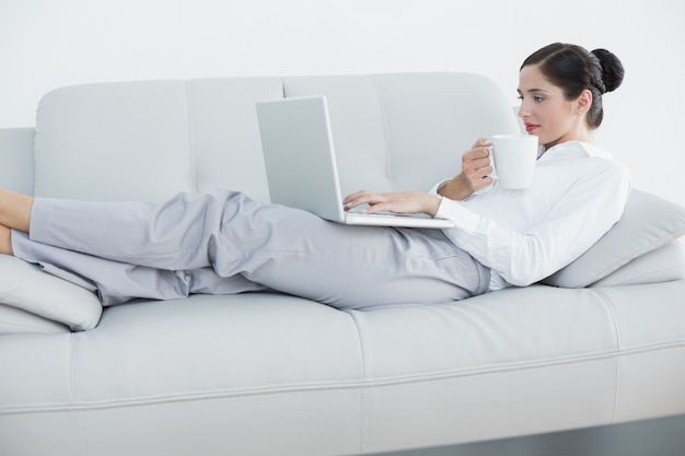 Gut gekleidete Frau mit Laptop und Kaffeetasse auf Sofa