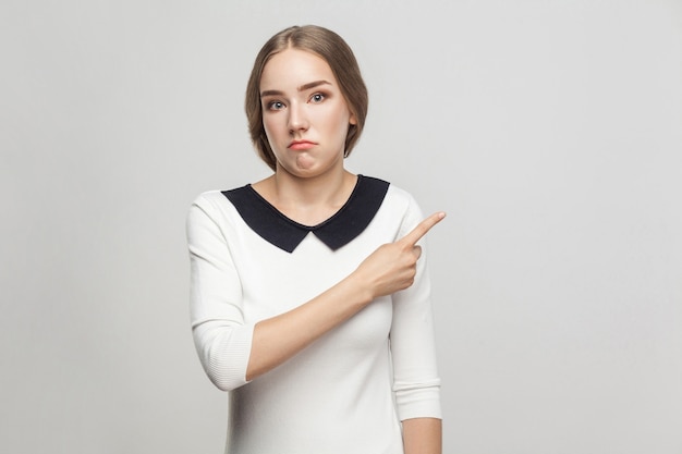 Gut gekleidete schöne Frau mit gesammelten Haaren, die mit verwirrtem Gesicht mit dem Finger auf den Kopierraum zeigen