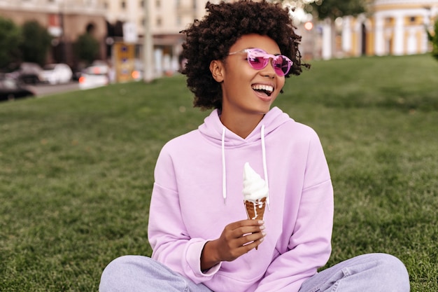 Gut gelauntes attraktives Mädchen in rosa Sonnenbrille, lila Hoodie und Jeans lächelt, sitzt draußen auf Gras und hält Eis