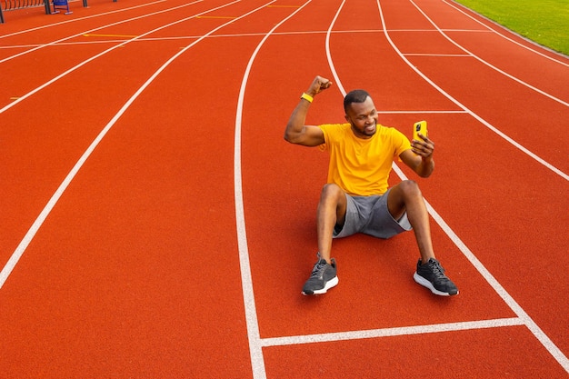 Gutaussehender afrikanisch-amerikanischer Mann in gelbem T-Shirt, der lächelt und die geballte Faust hebt, die Muskeln anspannt und ein Foto auf dem Smartphone macht Glücklicher männlicher Trainer sitzt im Stadion und macht eine Pause vom Training