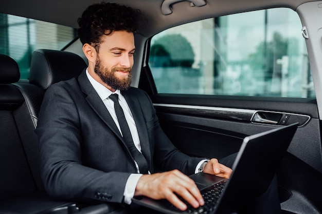 Gutaussehender, bärtiger, lächelnder Manager, der auf dem Rücksitz des Autos an seinem Laptop arbeitet